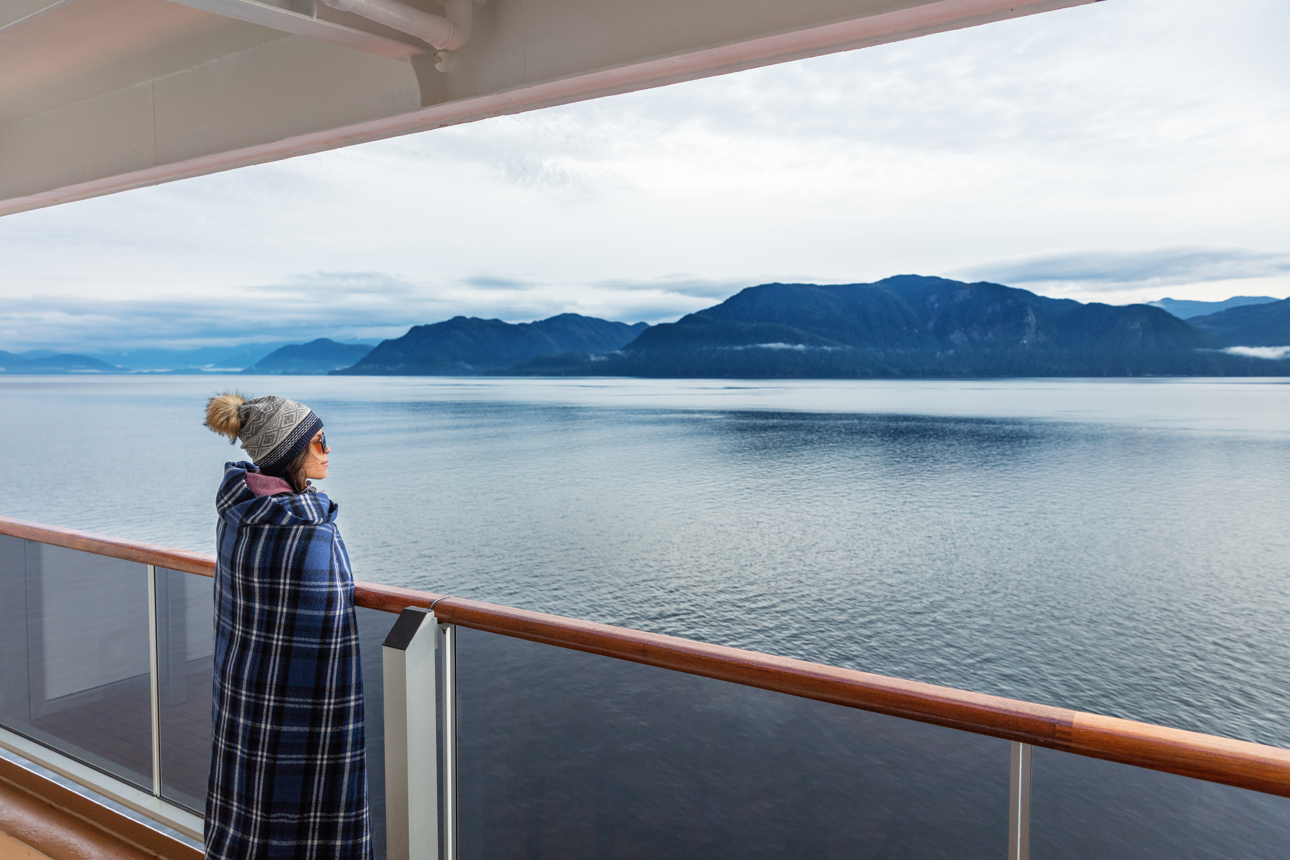 Alaska Cruise Travel Luxury Vacation Woman Watching inside Passage Scenic Cruising Day on Balcony Deck Enjoying View of Mountains and Nature Landscape. Asian Girl Tourist with Wool Blanket.